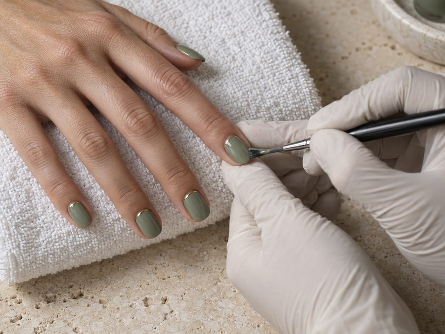 Artist painting sage-green gel polish with a thin gold cuticle line